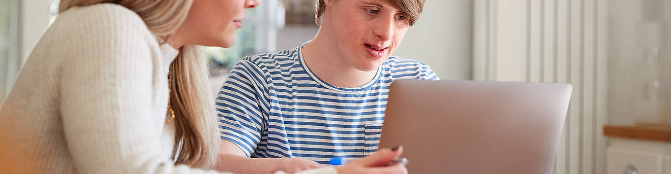 Zwei Personen sitzen an einem Tich. Eine Person hat ein aufgeklapptes Laptop vor sich stehen.